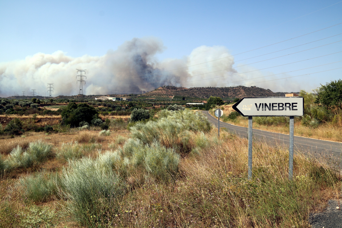 Un senyal de trànsit indica la localitat de Vinebre amb un gran núvol de fum d'un incendi forestal al fons