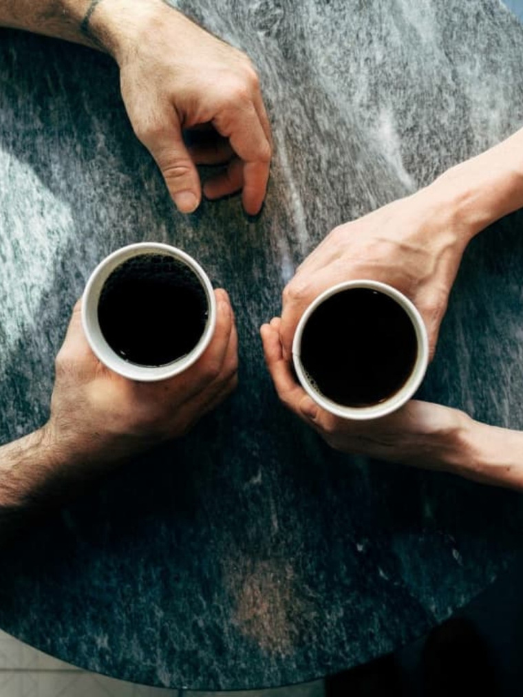 Imagen de dos personas sosteniendo dos tazas de café sobre una mesa