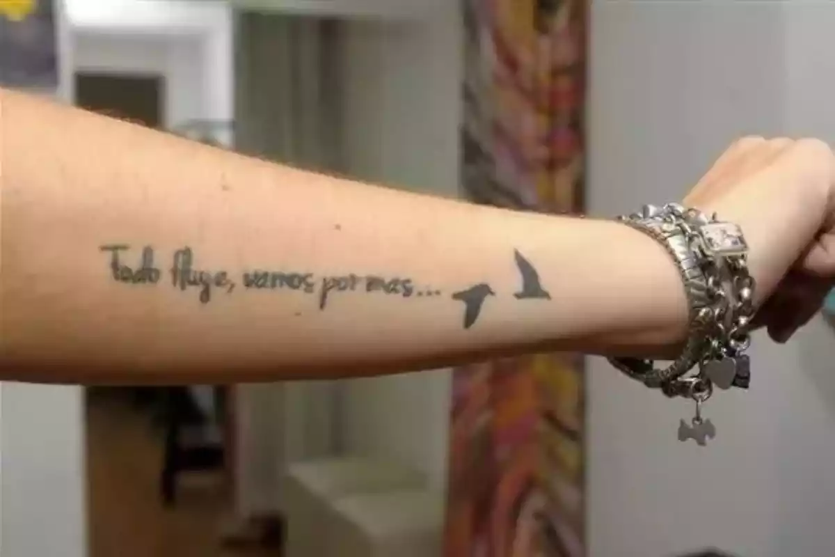 Foto de un brazo con reloj y pulseras que tiene además un tatuaje de una frase en negro: 'Todo fluye vamos por más....' Foto de un brazo con reloj y pulseras que tiene además un tatuaje de una frase en negro: 'Todo fluye vamos por más....'