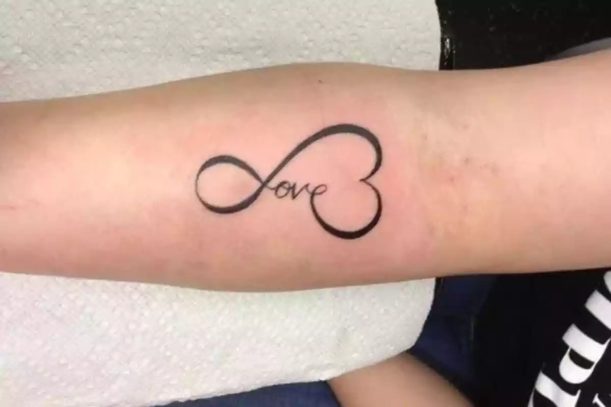 Foto de un brazo con un tatuaje en negro que forma el símbolo del infinito con un corazón y la palabra LOVE Foto de un brazo con un tatuaje en negro que forma el símbolo del infinito con un corazón y la palabra LOVE