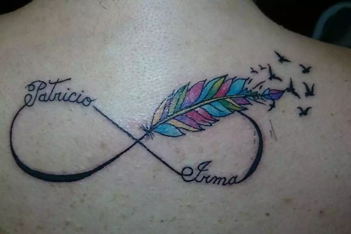 Foto de una espalda en la que aparece un tatuaje del símbolo del infinito con dos nombres y una pluma de colores que se deshace en pájaros Foto de una espalda en la que aparece un tatuaje del símbolo del infinito con dos nombres y una pluma de colores que se deshace en pájaros