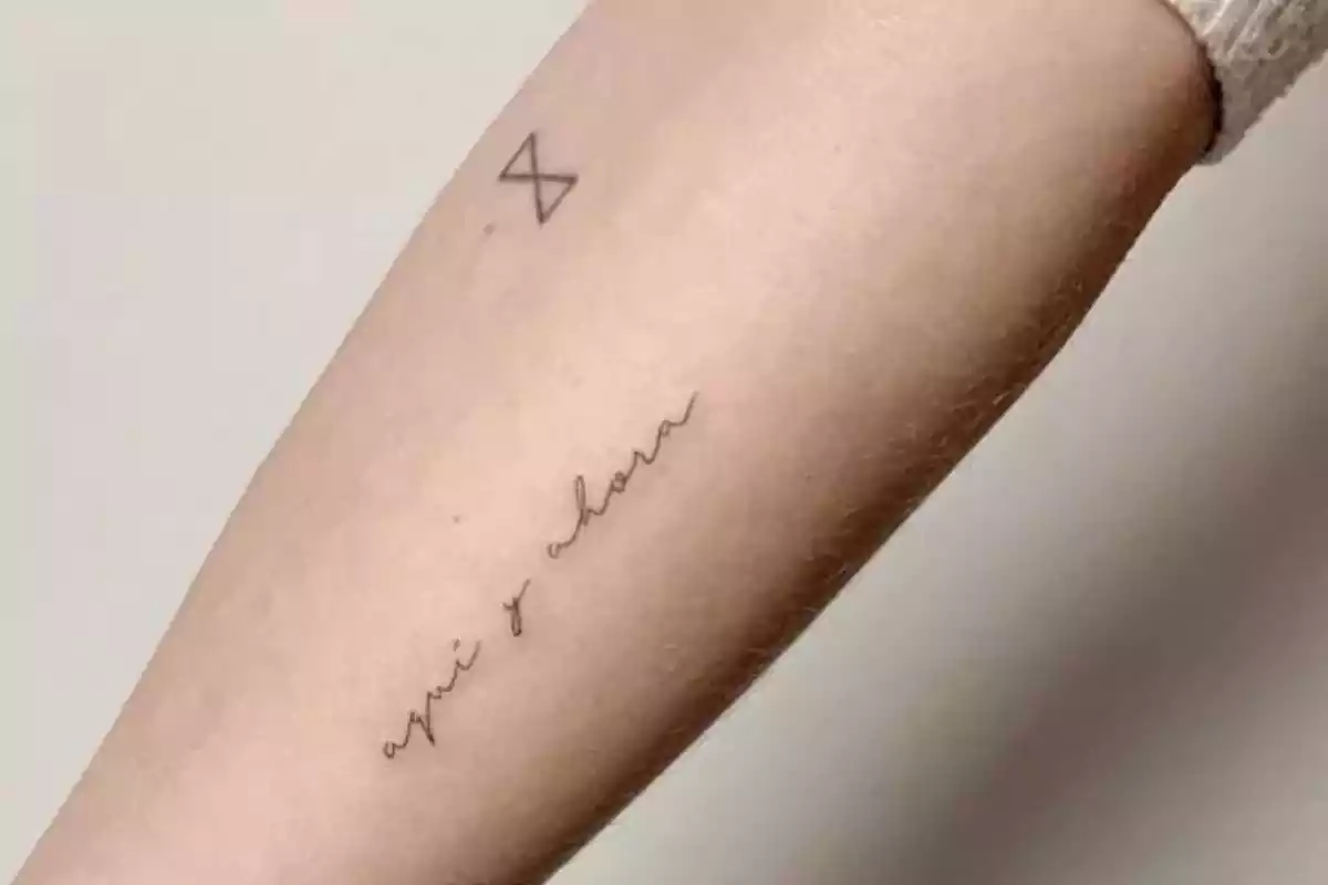 Foto recortada de un brazo en el que aparecen dos diseños de tatuajes. Uno de una X y otro con la frase 'Aquí y ahora' Foto recortada de un brazo en el que aparecen dos diseños de tatuajes. Uno de una X y otro con la frase 'Aquí y ahora'