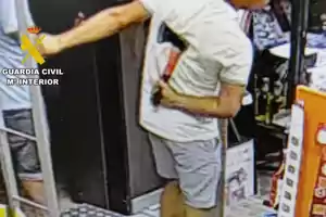 Persona con camiseta clara y pantalón corto sosteniendo un hacha dentro de una tienda con el logo de la Guardia Civil en la esquina superior izquierda