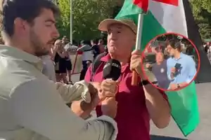 Un hombre sostiene una bandera y un micrófono mientras discute con otra persona en la calle, al fondo hay más personas observando y en la esquina de la imagen aparece un recuadro con dos hombres, uno de ellos con micrófono y el otro gesticulando.