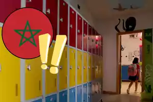Pasillo de escuela con casilleros de colores y una niña entrando a un aula, con un gráfico de la bandera de Marruecos y signos de exclamación superpuestos.