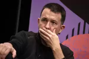 Un hombre con expresión pensativa se cubre la boca con la mano, vistiendo un suéter oscuro y con un fondo de colores púrpura y negro.